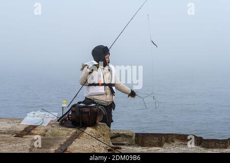 A winter picture of a woman with a fishing rod. Blue ocean and mist in the background. Picture from Malmo, Sweden Stock Photo