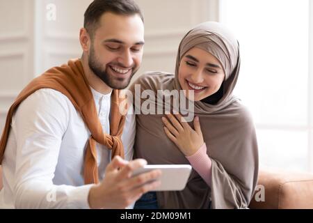 Muslim Couple Using Smartphone And Laughing Sitting In Living Room Stock Photo