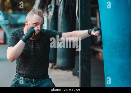 Street Fighter in Black Clothes and Bandages on the Wrist Boxing in Punching Bag Outdoors. Young Man Doing Box Training and Practicing His Punches at the Outside Gym Stock Photo