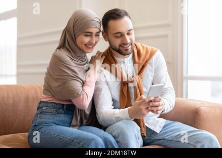 Young Muslim Couple Using Cellphone Sitting On Couch Indoors Stock Photo