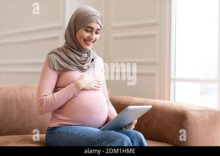 Joyful Pregnant Muslim Lady Using Digital Tablet Sitting At Home Stock Photo