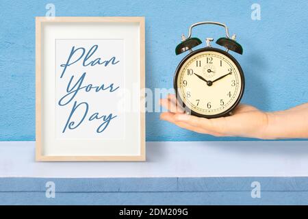 Hand holding an alarm clock next to a wooden frame with the sentence "Plan your time" as time management concept. Stock Photo