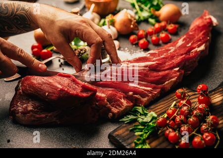 Fresh meat preparation and portioning.Whole uncut beef tenderloin.Raw meat seasoning.Man hands cutting up filet mignon steaks. Spices and vegetables f Stock Photo