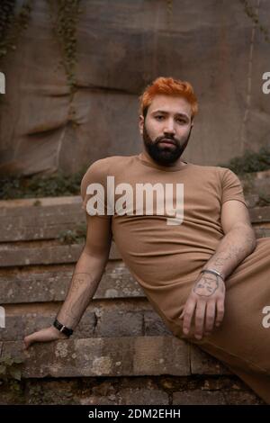 Portrait of young man with orange hair and brown dress sitting outdoor on stone stairs Stock Photo