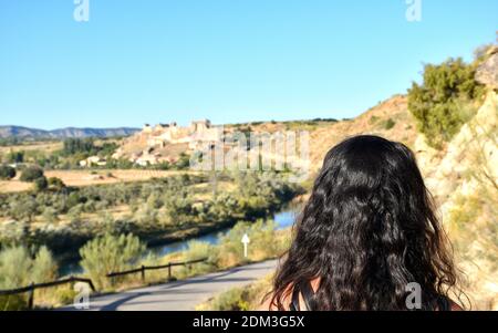 Long-haired dark-haired tourist observing the town of Zorita de los Canes and its castle from a distance. Tajo river between the two. Stock Photo