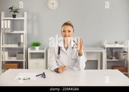 Young smiling blonde woman doctor nurse therapist sitting and greeting patient with hand online Stock Photo