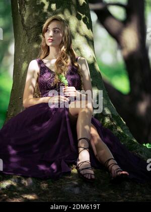 Naturally beautiful young women with long blonde hair dressed in a purple ball gown sits against a tree in the countryside in a dreamy romantic pose Stock Photo