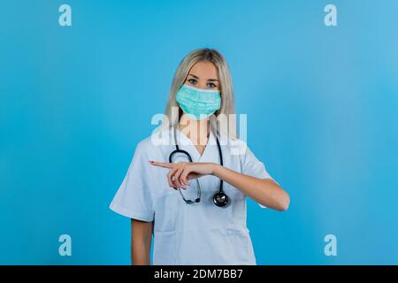 Female doctor with mask indicates a point. Portrait of a doctor in lab coat with face mask pointing to side at copy space isolated on light blue backg Stock Photo