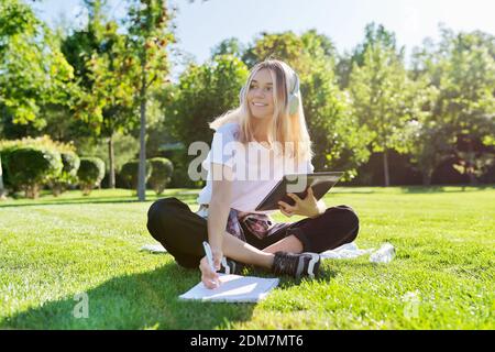 Teenage student in headphones with digital tablet sitting on lawn writing in notebook Stock Photo