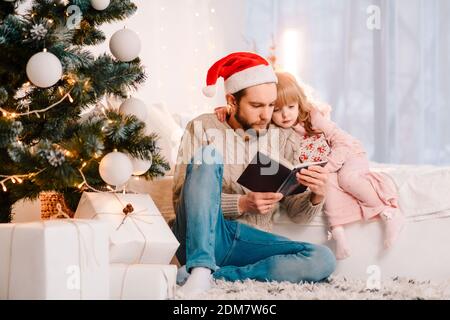 Dad in santa hat reading book with his daughter. Father reading fairy tale to a child Stock Photo