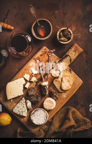 Snacks and appetizers variety for red wine concept. Flat-lay of various cheeses, crackers, smoked meat and glass of red wine on wooden rustic board over rusty brown background, top view Stock Photo
