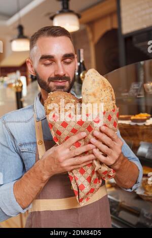 Vertical shot of a handsome male baker smelling freshly baked bread he is selling Stock Photo