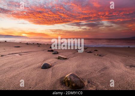 A Vibrant Bright Red Ocean Sunset With Beach Rocks in the Foreground Stock Photo