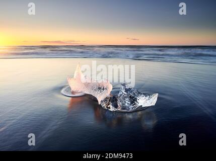 Clear majestic ice chunk cast on wet sandy beach against ocean in sunset Stock Photo