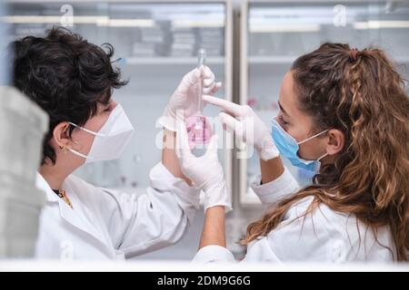 Two young female scientists wearing face masks using a flask. Laboratory research concept. Stock Photo