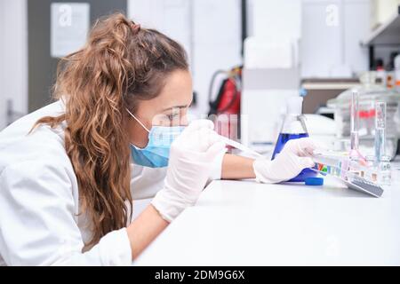 Young female scientist wearing face mask using a Pasteur pipette and a rack with tubes. Laboratory research concept. Stock Photo