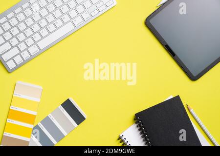 Graphic designer desk top view with tablet, color guide palette, keyboard, paper notebooks on yellow background. Creative professional workspace, view Stock Photo