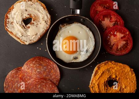 A flat lay breakfast or snack on black wood for one person: a sesame bagel toasted and sliced in half with  cream cheese on, slices of tomat,  sunny s Stock Photo