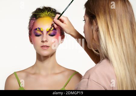 Female portrait with unusual face art makeup. Paint on brows, hair and around eyes. Artist's hand with paintbrush painting beautiful girl's brows make Stock Photo