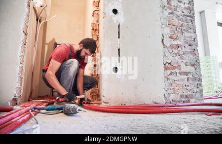 Man plumber in work overalls using hammer while laying pipes on the floor. Bearded male worker in gloves installing heating system while working on renovation of apartment. Concept of building job Stock Photo