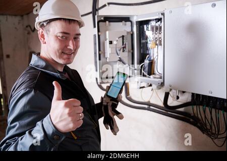 Portrait of smiling worker, wearing uniform and helmet, showing thumb up, standing near transformer, holding smartphone, making settings. Concept of working process Stock Photo