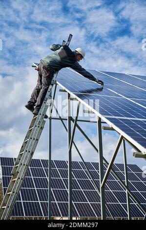 Vertical snapshot of working process in a solar field. Worker in protective uniform, standing on a ladder trying to set solar batteries on metal ground mounting. Concept of renewable energy resource Stock Photo