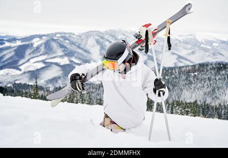 Alpine skier in snowboard jacket holding skis and ski poles while trying to walk through deep white snow. Man spending vacation in mountains. Concept of winter sport activities. Stock Photo