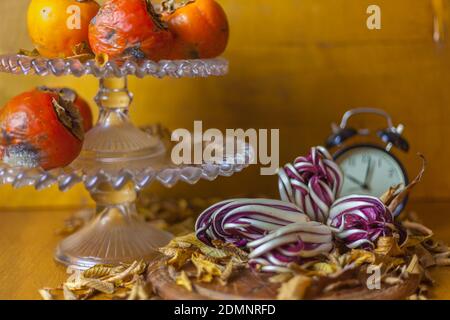Fruit holder with persimmon and red radicchio on wooden table with a base of dry yellow leaves. Presence of blurred old alarm clock on the right side Stock Photo