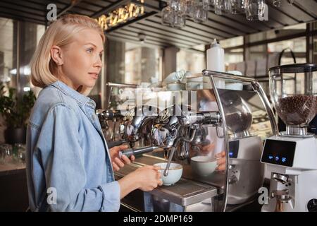 Attractive female barista looking away thoughtfully while making coffee for a customer, using coffee machine, copy space Stock Photo