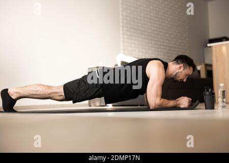 Plank it Training at home. Confident young man wearing black sportswear and doing plank position while exercising on the floor in light loft interior. Stock Photo