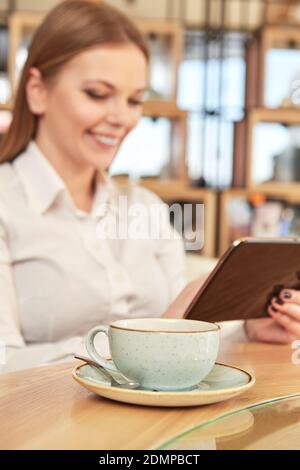 Web surfing. Selective focus on the cup on the cafe table cheerful woman using digital tablet on the background Stock Photo
