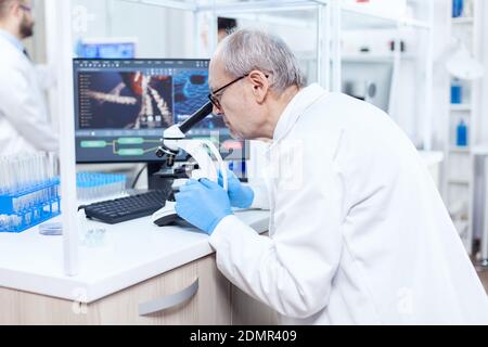 Senior caucasian scientist microbiology laboratory conducting virus analysis looking through microscope. Chemist researcher in sterile lab doing experiments for medical industry using modern technology. Stock Photo