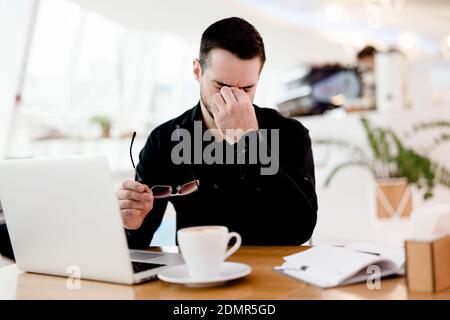 Give your eyes a rest Young tired freelancer man in black shirt suffers from pain and dryness in the eyes. He works a lot Cozy coffeehouse atmosphere Stock Photo