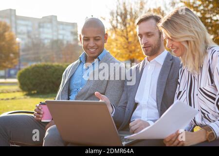 Diverse group of business colleagues using laptop outdoors. Business team meeting at the park, using computer together Stock Photo