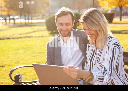 Two cheerful business colleagues enjoying working outdoors on a warm autumn day, using laptop together. Entrepreneurship, startup concept Stock Photo