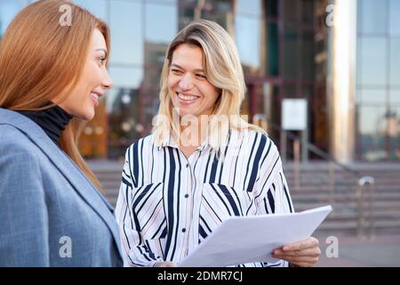 Charming mature businesswoman discussing startup with her colleague. Female entrepreneurs talking outdoors in front of office building Stock Photo