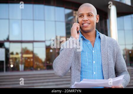 Handsome cheerful African American businessman smiling, talking on the phone outdoors. Happy young male entrepreneur calling someone on the phone Stock Photo