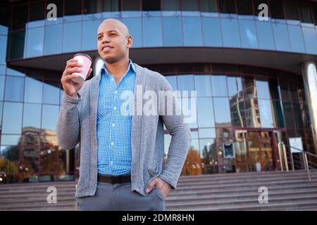 Low angle shot of a happy handsome African man having coffee break outdoors, office building on background Stock Photo