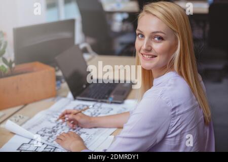 Achievement, business strategy concept. Happy beautiful female architect smiling to the camera over her shoulder, sketching construction plan at her o Stock Photo