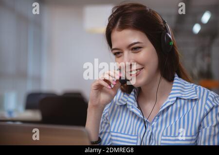 Close up of a beautiful friendly female customer support operator working at the call center, using laptop. Charming young woman wearing headset with Stock Photo