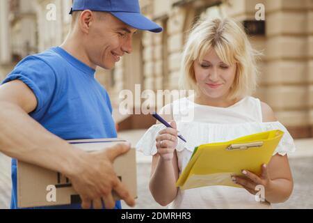 Cheerful woman receiving cardboard box parcel from a friendly male courier. Happy deliveryman smiling, holding post box while client signing papers on Stock Photo