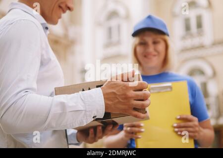 Cropped close up of a man holding cardboard box, happy female courier smiling on background. Satisfied customer smiling, holding parcel post box deliv Stock Photo