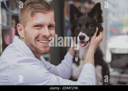 Handsome happy male veterinarian laughing to the camera, petting adorable dog at his clinic. Cute black puppy being examined by professional vet docto Stock Photo