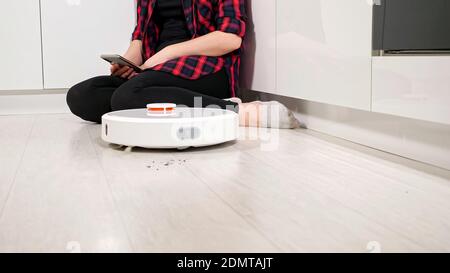 Unrecognizable woman with phone sits on the floor while the robot vacuum cleaner cleans the floor. Stock Photo