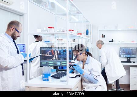 Assistant doing research using microscope and scientist taking notes on tablet pc. Team of researchers doing pharmacology engineering in sterile laboratory for healthcare industry with african assistant in the background. Stock Photo
