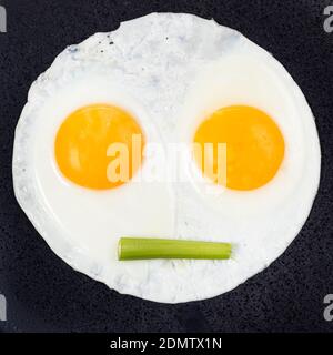 fried eggs and piece of celery on black plate close up. Fried eggs like face with straigh closed mouth Stock Photo