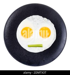 top view of fried eggs and piece of celery on black plate isolated on white background. Fried eggs like face with straigh closed mouth Stock Photo