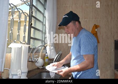 Elderly man washing dishes with sponge in kitchen sink at home, wearing T-shirt and cap in front of glass window, Brazil, South America Stock Photo