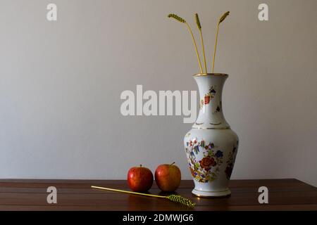 Floral pottery vase, apples and ears of wheat on wooden table on white wall background. Still life photo with empty space for text Stock Photo