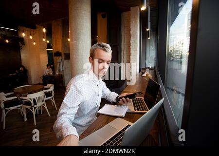 Freelance programmer sits near the window in a coffee shop and works. Stylish blonde man in white shirt uses laptops. Cozy coffee shop atmosphere. Caf Stock Photo